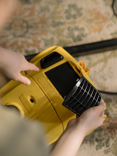 A person operating a yellow carpet cleaning machine with a black handle and vent, positioned on a patterned rug in a domestic setting. The cleaning device is in use, with the person's hands guiding it
