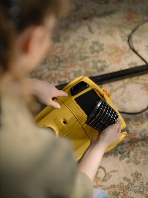 A person operating a yellow carpet cleaning machine with a black handle and vent, positioned on a patterned rug in a domestic setting. The cleaning device is in use, with the person's hands guiding it