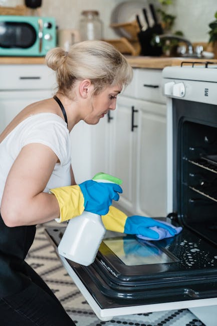 A woman with blond hair tied back, wearing a white t-shirt and yellow and blue cleaning gloves, is engaged in surface cleaning inside a kitchen. She is holding a white spray bottle and is wiping the i